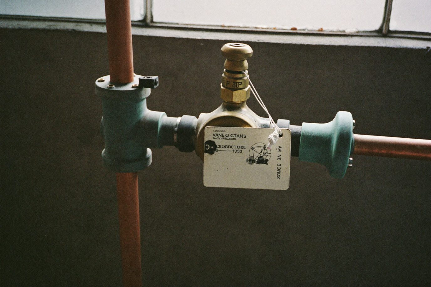 Brass backflow preventer assembly on copper pipe with a worn metal inspection tag wired to it, in a dim utility room