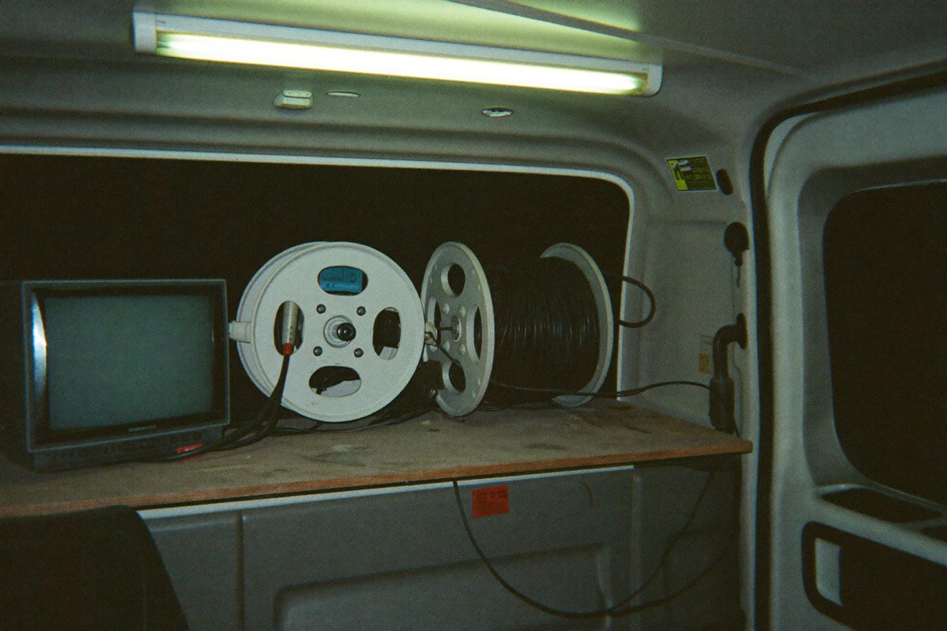 Sewer inspection camera crawler and cable reel on a plywood shelf in a work van beside a small field monitor