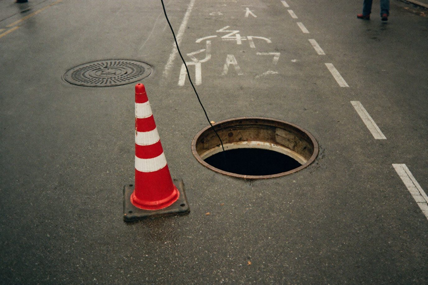 Open manhole with a safety cone on wet asphalt and a camera cable running down into the opening