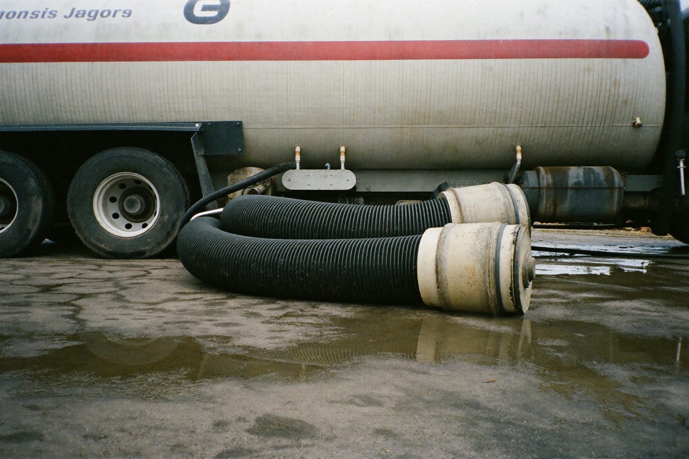 Heavy suction hose from a vacuum tanker lying across wet stained concrete, truck out of focus behind