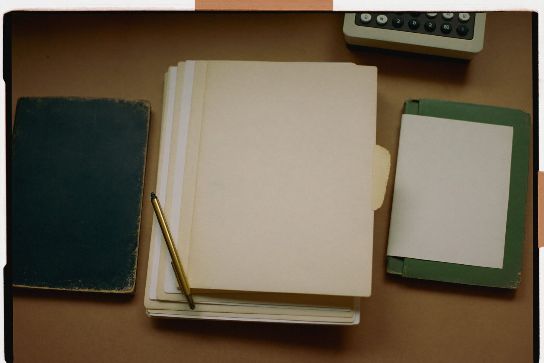 Overhead photograph of closed manila folders, a worn ledger book, a brass pencil and an adding machine on a desk